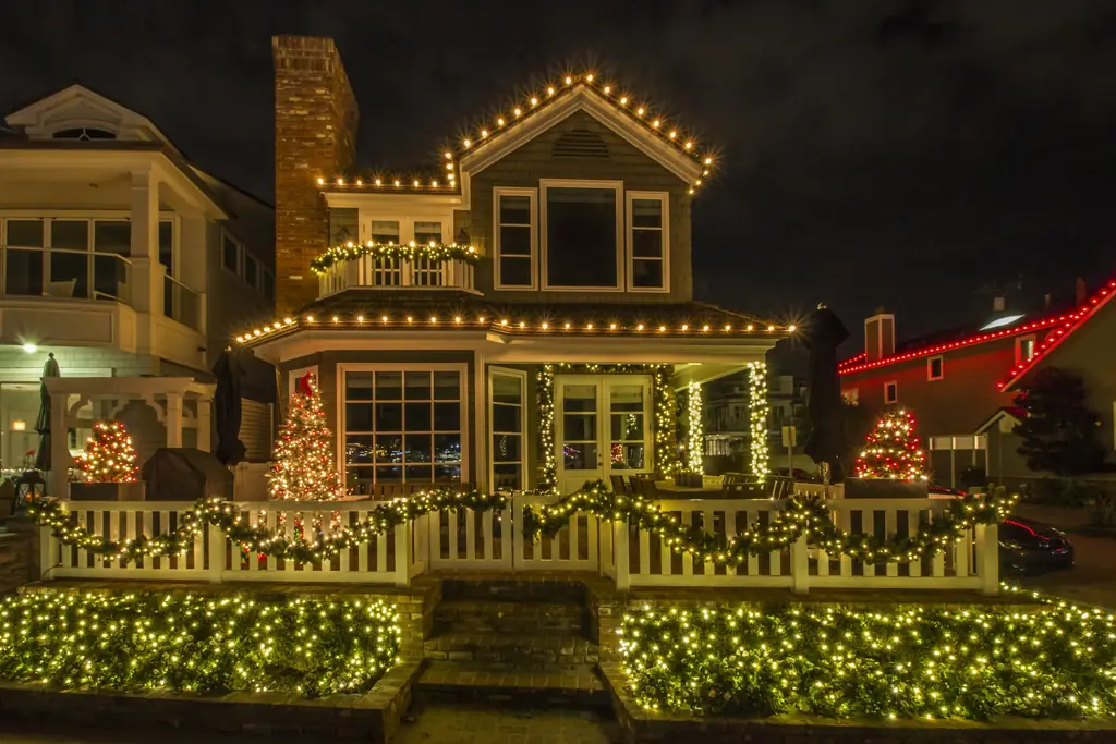 Two-story home with warm white roofline lights, lit shrubs, and garland on the fence