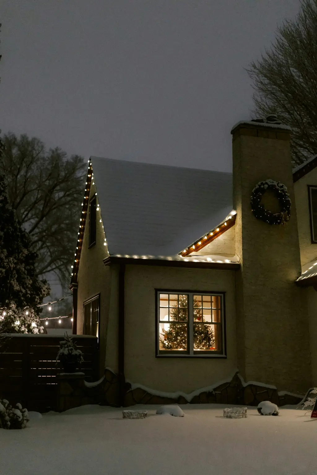Snow-covered home with a lit roofline and a wreath glowing beside the window